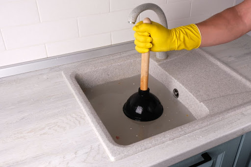 Homeowner wearing yellow glove holding a plunger over clogged kitchen sink