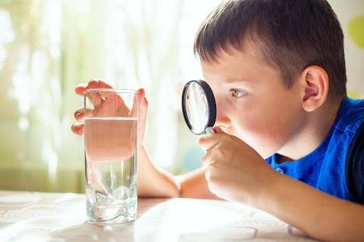 Kid holding a magnifying glass is inspecting water in a glass.