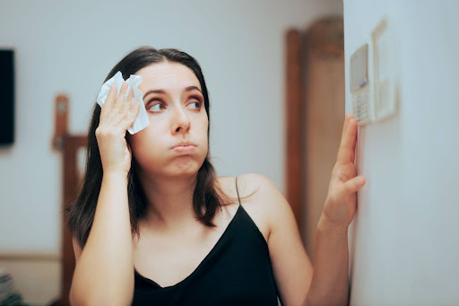 Woman wiping sweat from her brow due to broken AC while looking at thermostat.